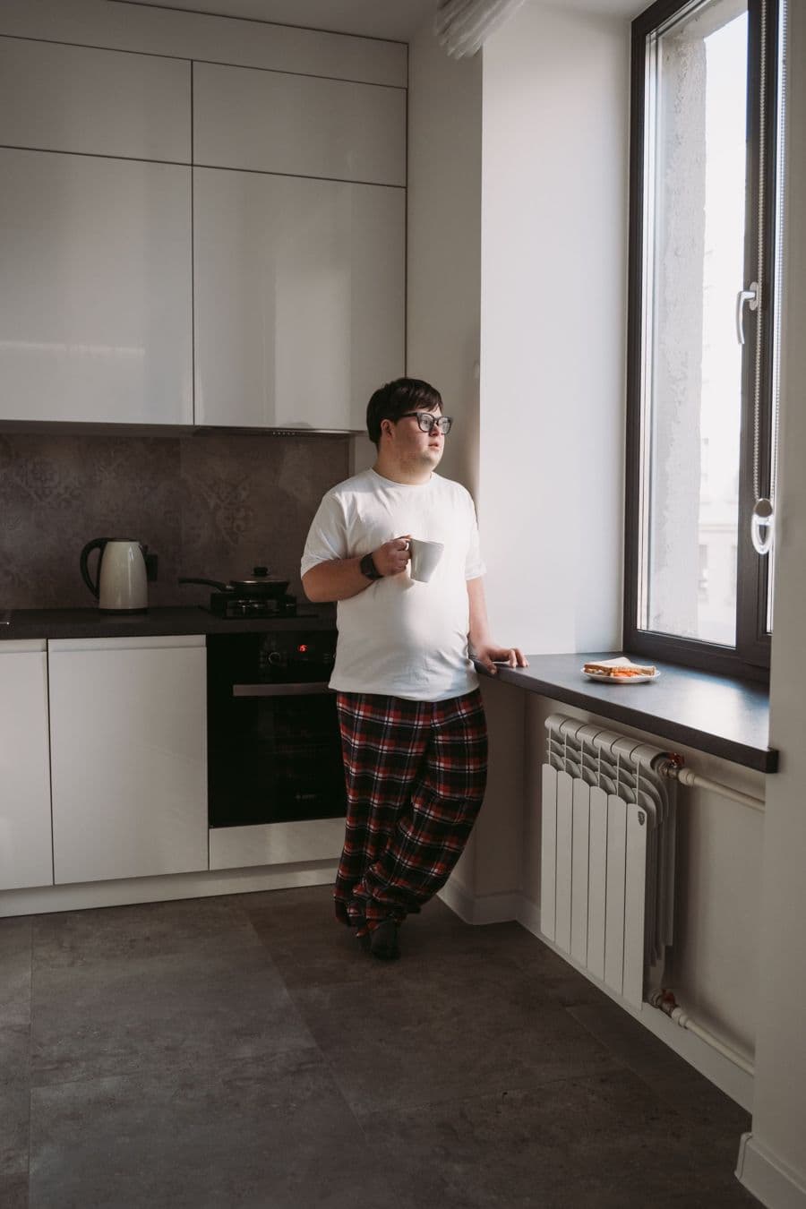 A young man with Down syndrome in his own kitchen, holding a mug by the window